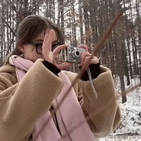 Девушка поднимает камеру, делает вид, что снимает, опускает её, делает игривый жест рукой у губ и снова поднимает камеру. — девушка в бежево