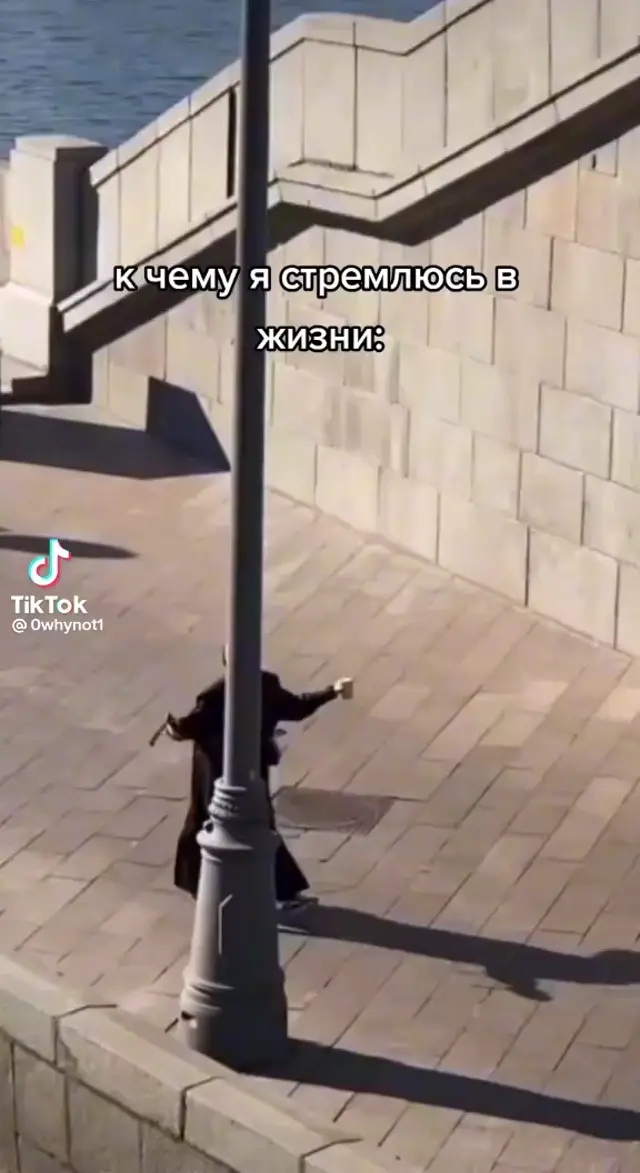 Человек в черном длинном пальте пританцовывает и припрыгивает, идя по пустой набережной под музыку. — человек в черном пальте