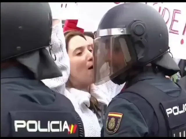 Протестующая кричит на полицейского в упор, а затем с улыбкой приближает губы к его каске, имитируя поцелуй, после чего держит плакат высоко