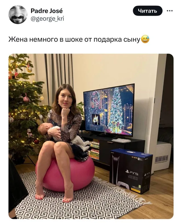 Женщина сидит на фитболе, держит на руках спящего младенца и смотрит в камеру с скептическим выражением лица, рядом стоит коробка от PS5 — ж