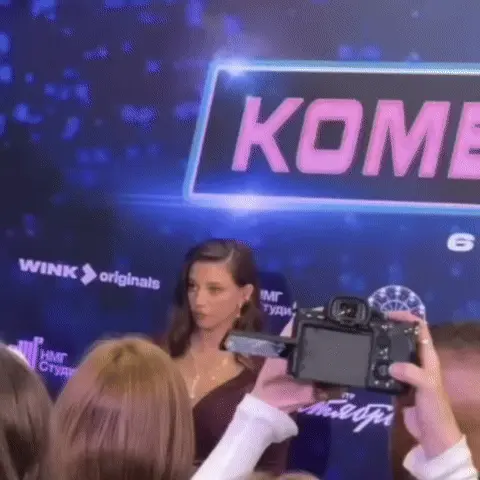 Девушка стоит неподвижно, пока вокруг неё суетятся фотографы, периодически слегка поворачивая голову. — девушка с длинными темными волосами 