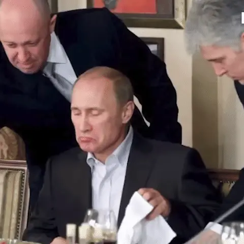 Евгений Пригожин склоняется над сидящим Владимиром Путиным, который сохраняет неподвижную позу с поджатыми губами. — Владимир Путин, Евгений