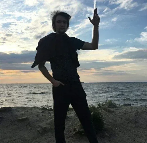 Парень стоит в расслабленной позе, подняв руку в жесте «пальцевого пистолета» на фоне неба. — молодой человек с растрепанными волосами