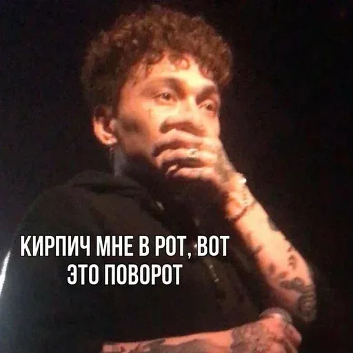 Человек стоит в полуобороте, прикрыв рот рукой, словно услышал что-то невероятное — мужчина с кудрявыми волосами и татуировками