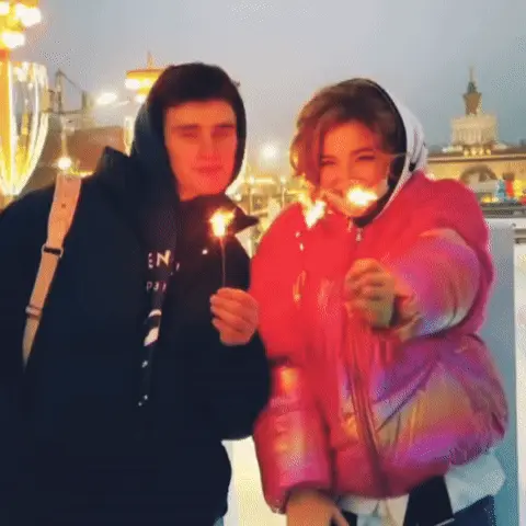 Молодые люди стоят рядом, держат бенгальские огни, которые активно искрят, создавая световые блики на их лицах. — парень в черной толстовке,