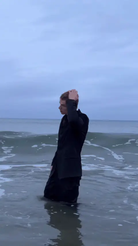 Парень стоит в морской воде, волна окатывает его с головой, после чего он стряхивает воду, поправляет галстук и готовится к следующей волне.