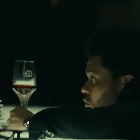 The Weeknd сидит неподвижно, слегка поворачивая голову и переводя взгляд, при этом бокал вина остается в кадре как часть композиции. — The W