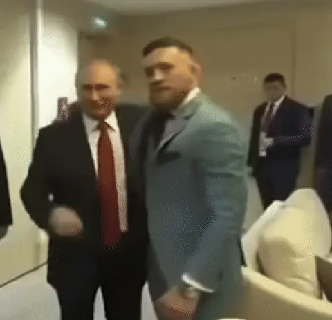 Мужчина в сером костюме приобнимает собеседника, затем делает резкое движение рукой, имитирующее удар или жест «силы», при этом его лицо ост