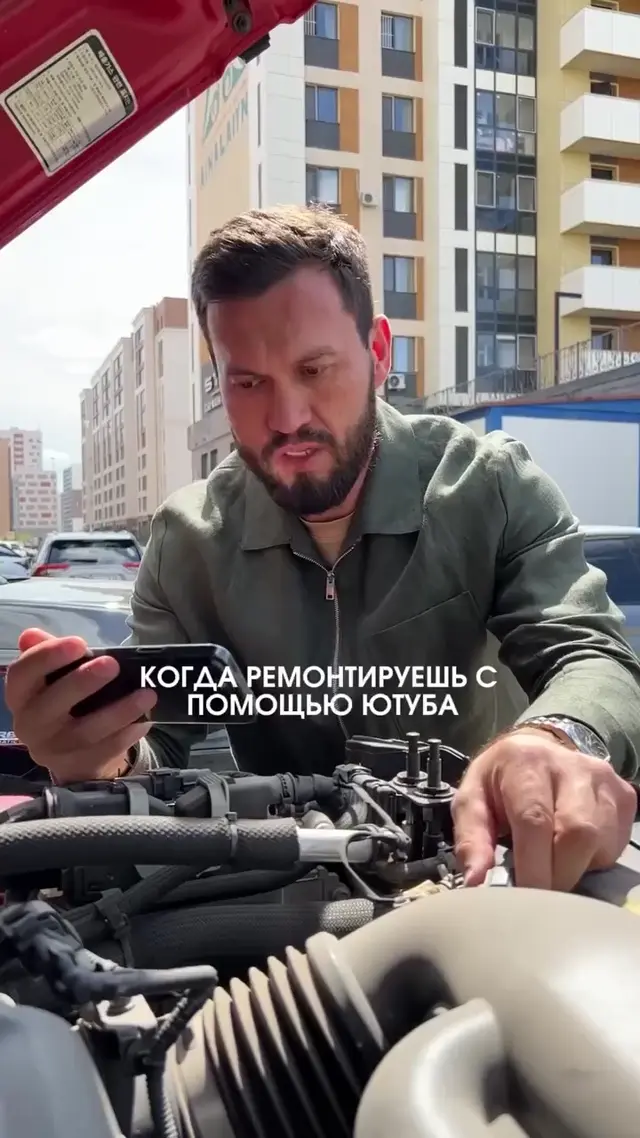 Мужчина смотрит в телефон, читая или слушая инструкцию, и одновременно пытается манипулировать проводами в моторном отсеке, периодически мен