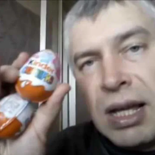 Мужчина держит два яйца «Kinder Сюрприз» на уровне лица, демонстрируя их зрителю — мужчина с седыми волосами