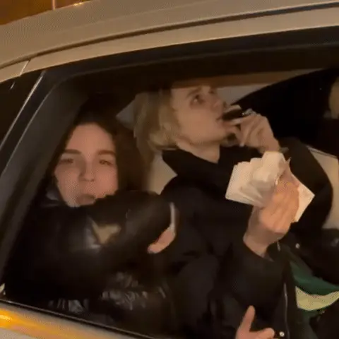 Парни сидят в машине, один из них периодически подносит вейп ко рту, другой слегка поправляет пачку денег в руках, оба смотрят в окно/на кам